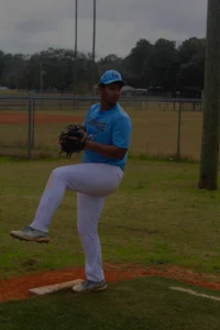 A baseball player in a light blue jersey and white pants is preparing to pitch on a grassy field with a baseball diamond in the background.
