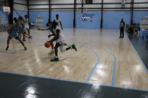 A basketball player dribbles the ball while being closely defended by opponents on a wooden court, with a referee observing the play