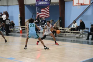 A basketball player dribbles while guarded by an opponent during a game, with spectators and an American flag in the background