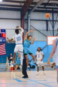 A basketball player in a white jersey leaps for a jump shot, while other players in blue and white compete for the ball in a gym