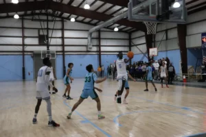 A basketball player jumps to shoot while teammates and opponents watch in an indoor gym, with a score displayed in the background
