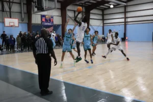A basketball player leaps for a shot while teammates and opponents watch, with the scoreboard showing 65-57 and a referee observing