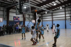A basketball player leaps to dunk while defenders jump to block, with spectators and scoreboard in the background during an intense game