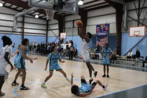 A basketball player leaps to make a shot while an opponent falls on the court; spectators cheer in the background. Scoreboard shows 51-41