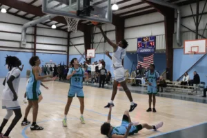 A basketball player leaps to make a shot while an opponent falls to the ground, with spectators and a scoreboard in the background