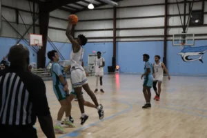 A basketball player leaps to shoot as opponents guard him on a well-lit court, surrounded by teammates and a referee in the foreground