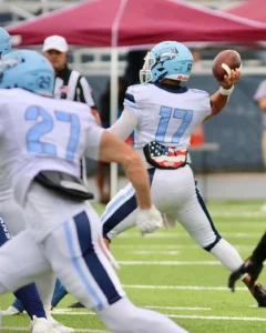 A football player in a light blue uniform prepares to pass the ball during a game, with teammates and a referee in the background.