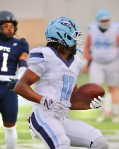 A football player in a white jersey with blue accents holds a brown football, poised to make a play on the field
