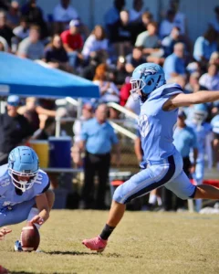 A football player prepares to kick, while another player holds the ball. A lively crowd watches in the background at an outdoor game