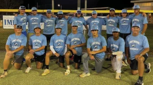 A group of 12 baseball players in light blue _Lighthouse Baseball_ shirts poses on a field at night, with a fence in the background