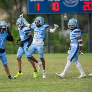A group of football players in blue jerseys celebrate on the field, with a scoreboard displaying a score of 10-1 in the background