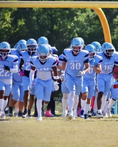 A group of high school football players in light blue uniforms run together on the field, showing teamwork and determination