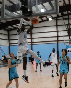 A player in a white jersey leaps to dunk a basketball, while opponents in teal jerseys watch on during an indoor game