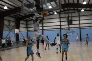 A player in a white jersey leaps to dunk a basketball while opposing team members watch in a gym with a wooden floor and bleachers