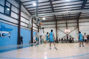 A player leaps for a slam dunk during a basketball game, while teammates and opponents watch in a gym decorated with a shark mural