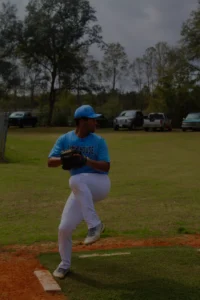 A young baseball player in a light blue uniform prepares to pitch on a grassy field, with vehicles parked in the background