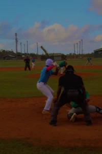 A youth baseball game in progress, featuring players in colorful uniforms on a grass field with cloudy skies in the background