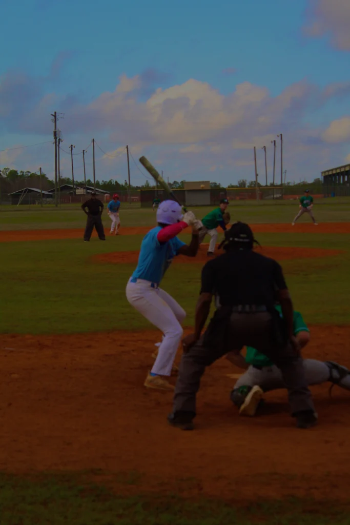 A youth baseball game in progress, featuring players in colorful uniforms on a grass field with cloudy skies in the background