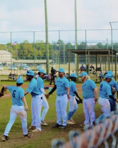 A youth baseball team in light blue uniforms celebrates a moment on the field, with players engaging and interacting enthusiastically.