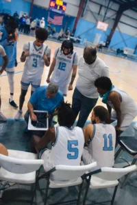 A youth basketball team huddles around a coach presenting strategies on a clipboard during a game in a gymnasium