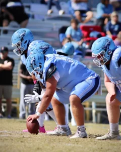 Three football players in light blue uniforms prepare for a snap on the field, focused on the game ahead. Spectators are visible in the background