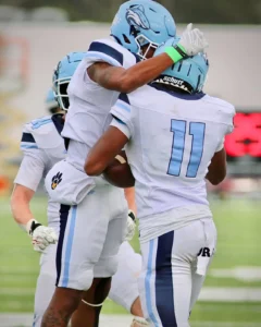 Two football players in light blue uniforms embrace in celebration on the field, with teammates visible in the background